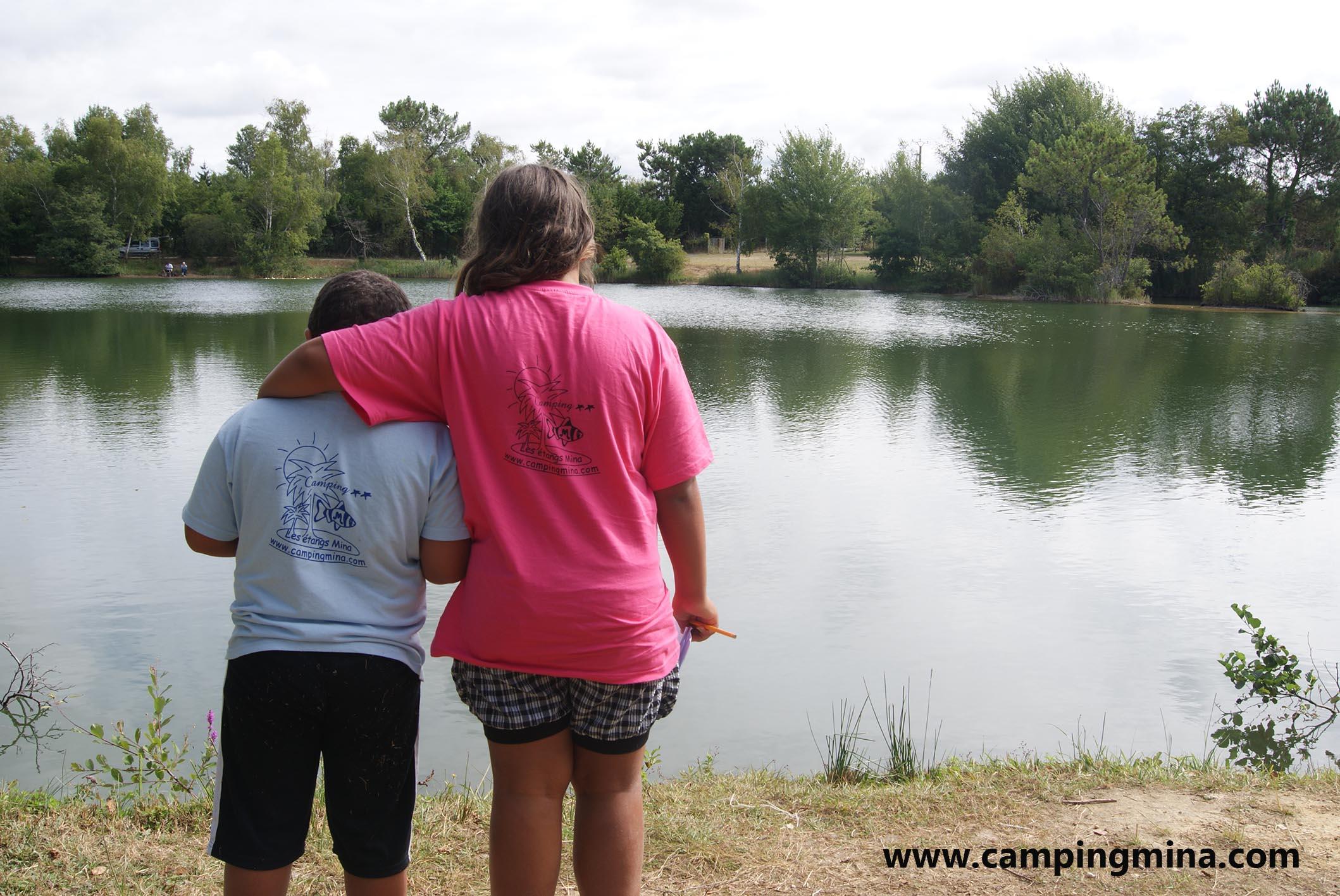 Zwei Kinder stehen am Seeufer und schauen auf das ruhige Wasser im Campingbereich, umgeben von Bäumen.