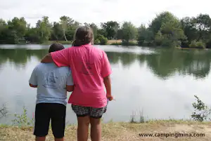 Zwei Kinder stehen am Seeufer und schauen auf das ruhige Wasser im Campingbereich, umgeben von Bäumen.