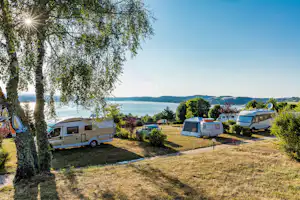 Wohnwagen und Zelte auf einem Campingplatz, im Hintergrund See und Bäume, daneben weitere Fahrzeuge.
