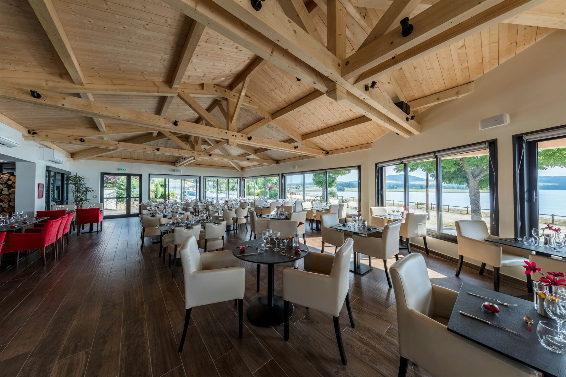 Innenraum eines Camping-Restaurants mit Holzbalkendecke, großen Fenstern und gedeckten Tischen mit Stühlen.