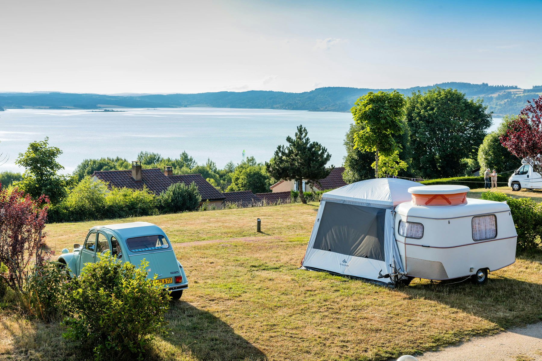 Zelt und Wohnwagen auf einer Campingwiese, im Hintergrund See und Hügel, daneben ein kleiner Transporter.
