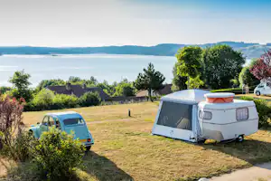 Zelt und Wohnwagen auf einer Campingwiese, im Hintergrund See und Hügel, daneben ein kleiner Transporter.