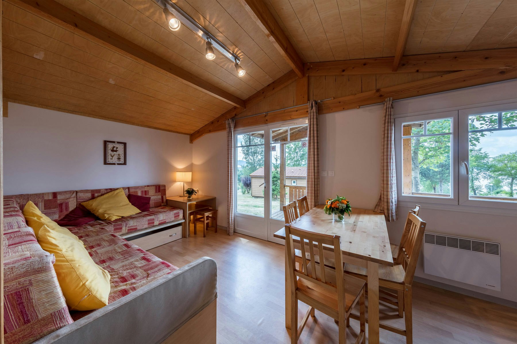 Innenansicht eines Camping-Unterkunftsraums mit Doppelbett, Holzbalkendecke, Esstisch und Stühlen sowie großen Fenstern zum Blick ins Grüne.