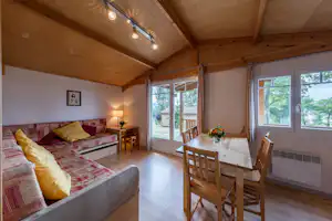 Innenansicht eines Camping-Unterkunftsraums mit Doppelbett, Holzbalkendecke, Esstisch und Stühlen sowie großen Fenstern zum Blick ins Grüne.