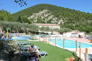 Poolbereich auf einem Campingplatz mit Liegen am grünen Hang, dahinter ein Gebäude und Bäume.