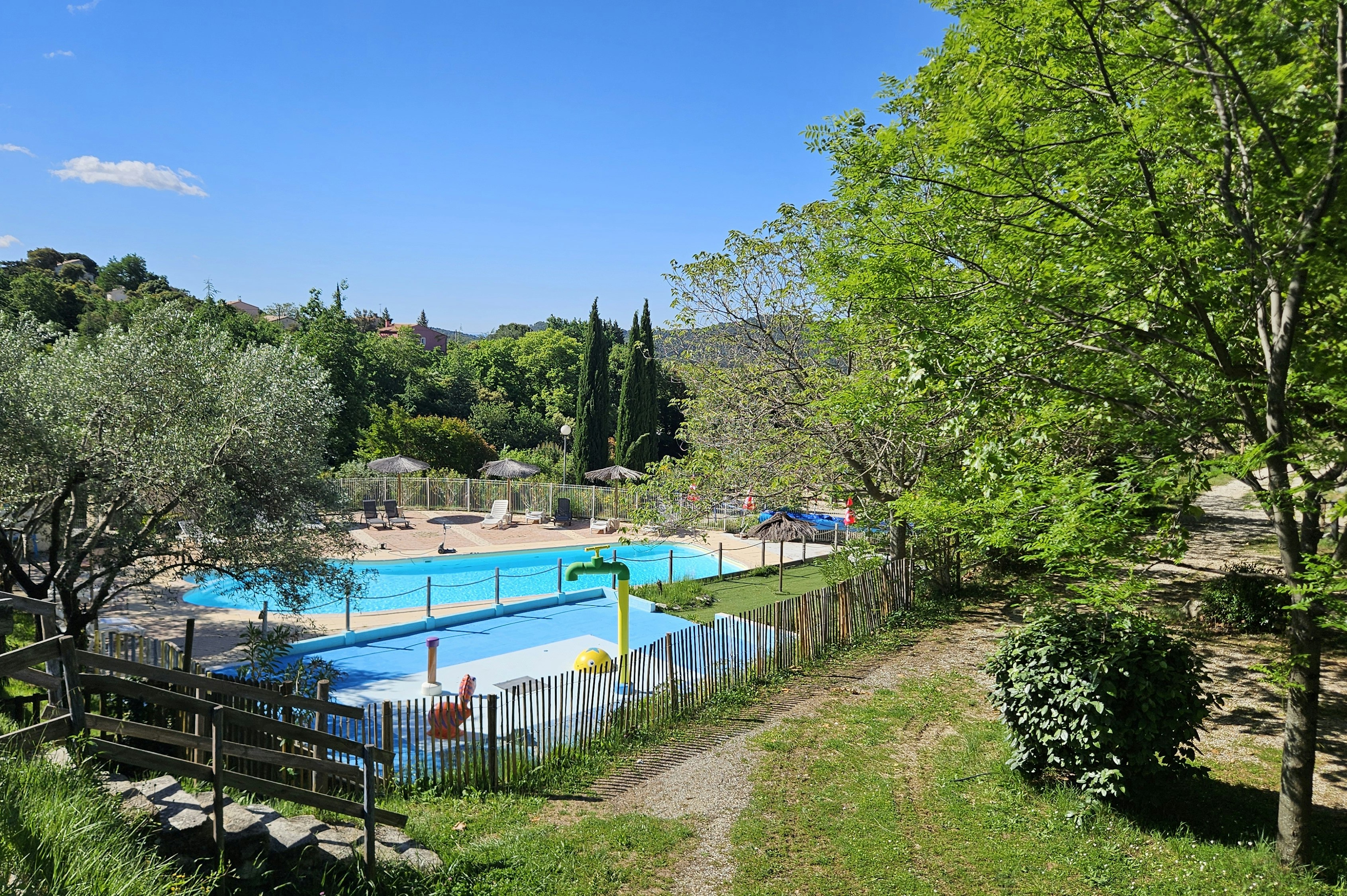 Schwimmbecken auf einem Campingplatz, eingezäunt mit Bäumen und Hecken, daneben ein Weg und Sitzbereich am Zaun.