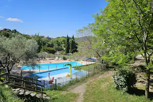 Schwimmbecken auf einem Campingplatz, eingezäunt mit Bäumen und Hecken, daneben ein Weg und Sitzbereich am Zaun.