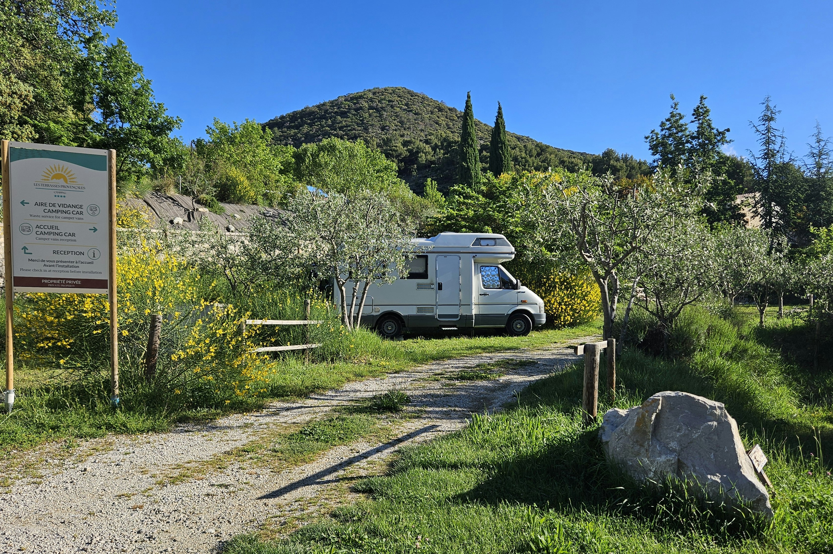 Wohnmobil auf Stellplatz am Campingplatz, Schotterweg, Hecken und Bäume, im Hintergrund Hügel und Zypressen.