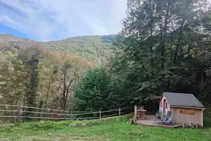 Kleine Campinghütte mit Holzterrasse am Hang, umgeben von Wiesen, Zaun und hohen Bäumen im grünen Tal.