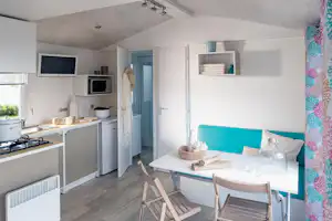 Innenansicht eines Wohnwagens mit Küchenzeile, Esstisch und blauer Sitzbank, Regalen und TV im hellen Raum.