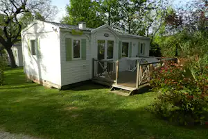 Mobilhome mit Holzterrasse und grünen Türen im Grünen, umgeben von Bäumen und Rasenfläche auf dem Campingplatz.