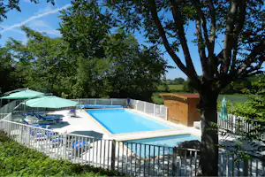 Camping-Pool mit blauer Wasserfläche, Liegen und Sonnenschirm, umgeben von Bäumen und weißem Zaun.