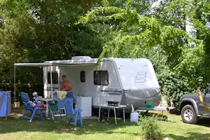 Wohnmobil mit Vorzelt und Campingmöbeln auf dem Stellplatz, daneben ein Auto und grünes Gras unter Bäumen.