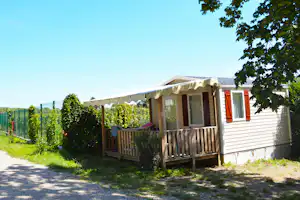 Mobilheim mit Holzterrasse, Markise und orangefarbenen Fensterläden, umgeben von Hecken und Zaun im Grünen.