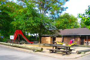 Spielplatz am Campingplatz: rote Rutsche, gelbe Wippe und Holzbänke vor Backsteingebäude, umgeben von Bäumen.