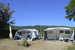 Zwei Wohnwagen auf dem Campingplatz mit Vorzelten, Campingstühlen und Tisch vor grünen Büschen und Hügeln.