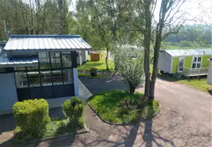Modernes Campinggebäude mit Glasfront, gepflastertem Weg und Rasenflächen, daneben ein Mobilheim und Bäume im Hintergrund.