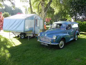 Blauer Oldtimer steht auf dem Campingplatz neben einem Zeltanhänger unter Bäumen auf grünem Rasen.
