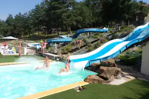Blaue Wasserrutsche am Campingplatz, Kinder rutschen über Treppen ins Schwimmbecken, im Hintergrund Bäume und Gebäude.