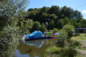 Aufblasbares Wasser-Spielgerät im Campingbereich: Kind springt von der blauen Rutsche über den Teich, im Hintergrund Bäume.