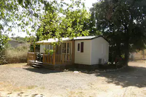 Mobilheim mit Holzterrasse, Stühlen und Tisch, umgeben von Bäumen und Hecken am Campingplatz.