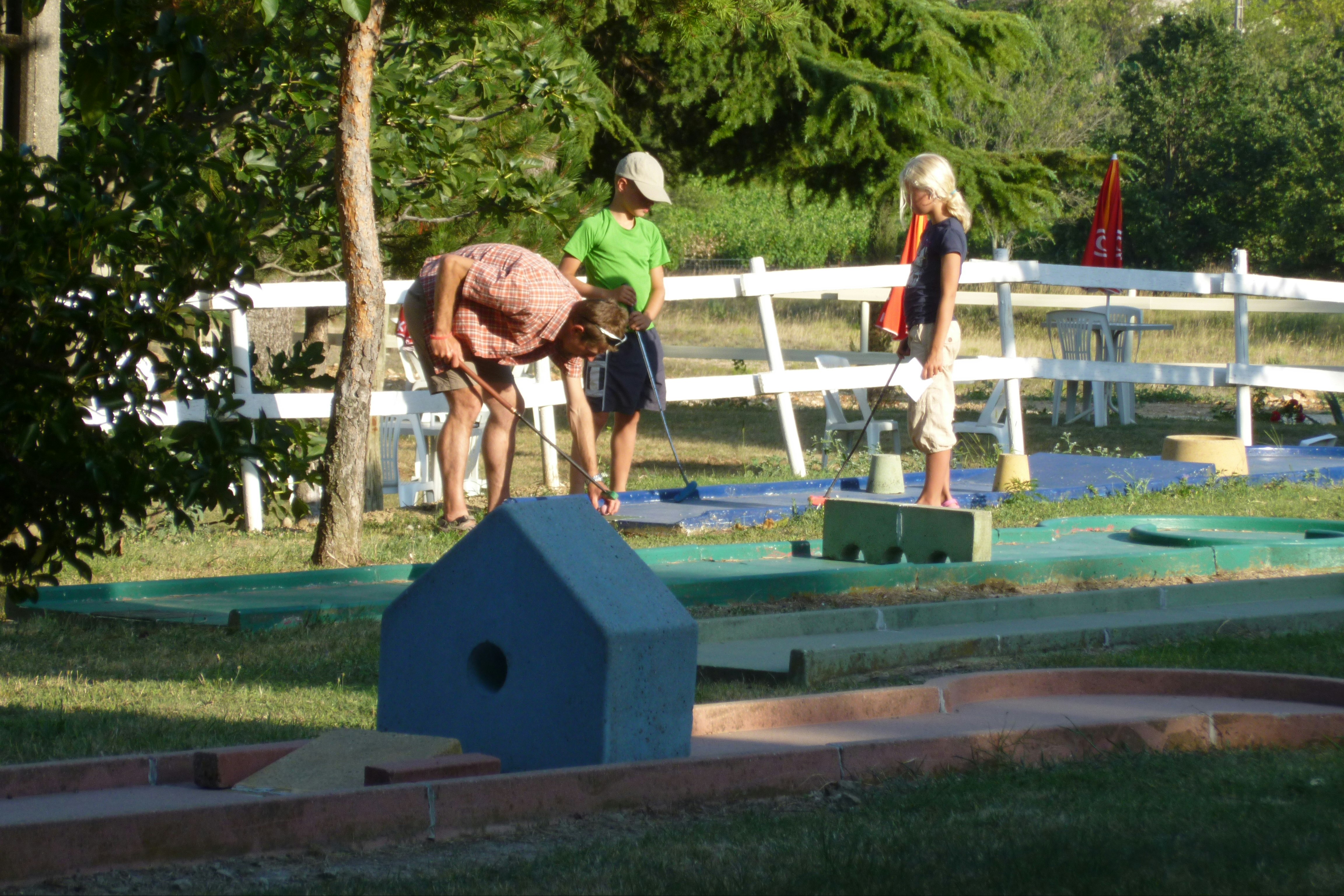 Familie beim Minigolf im Garten: Erwachsene und Kinder mit Schlägern an einem Bahnabschnitt, umgeben von Bäumen und Rasen.
