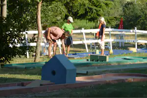 Familie beim Minigolf im Garten: Erwachsene und Kinder mit Schlägern an einem Bahnabschnitt, umgeben von Bäumen und Rasen.