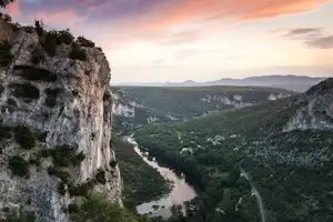 Blick auf einen Fluss im Tal, umgeben von bewaldeten Hängen und Felswand, mit Straße im Hintergrund.