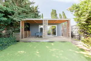 Modernes Holzhaus mit Terrasse, Sitzbereich und Rasenfläche, umgeben von Bäumen und Hecken im Campingbereich.