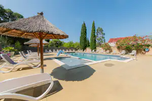 Poolbereich im Campingplatz: Sonnenschirme, Liegestühle und ein rechteckiges Becken mit Treppe, umgeben von Bäumen und Blumen.