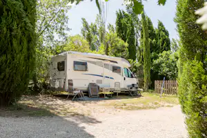 Wohnmobil auf einem Campingplatz-Stellplatz zwischen hohen Bäumen, mit Markise, Stühlen und Tisch vor dem Fahrzeug.