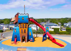 Buntes Spielhaus mit Rutsche auf einem Campingplatz, im Hintergrund Wohnwagen und Mobilheime sowie ein breiter Weg.