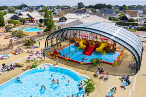 Überdachtes Schwimmbecken und großes Freibad auf dem Campingplatz, mit Liegestühlen, Wohnhäusern und Wegen im Hintergrund.