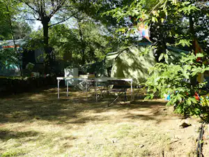 Zelt auf dem Campingplatz mit Tisch und Stühlen, umgeben von Bäumen und grünem Laub im Schatten.