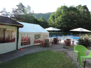 Zeltunterkünfte auf einer Holzterrasse am Campingplatz, mit Stühlen und Sonnenschirmen sowie Blick auf den Pool und Bäume im Hintergrund.