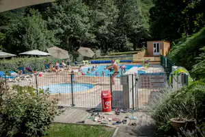 Camping-Pool mit blauem Wasser, Liegestühlen und Sonnenschirmen, eingezäunt mit Holzterrasse und kleinen Gebäuden im Hintergrund.
