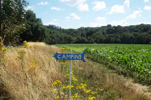 Camping-Schild an einem Feldweg, umgeben von Wiese, hohem Gras und Bäumen im Hintergrund.