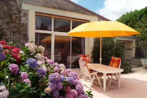 Terrasse am Campingplatz mit gelbem Sonnenschirm, weißem Tisch und Stühlen, Hortensien vor einem Steingebäude mit Fenstern.