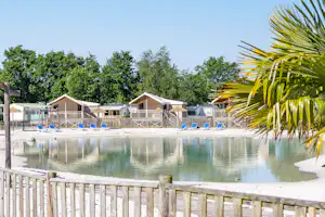 Schwimmbecken am Campingplatz mit Holzgeländer, Palmenzweig im Vordergrund und mehreren Bungalows im Hintergrund.
