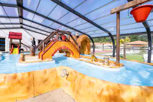 Innenbereich mit beheiztem Pool und Wasserrutsche, Steinbecken, Liegen und Glasdach, daneben Spielbereich mit Wasserfontäne.