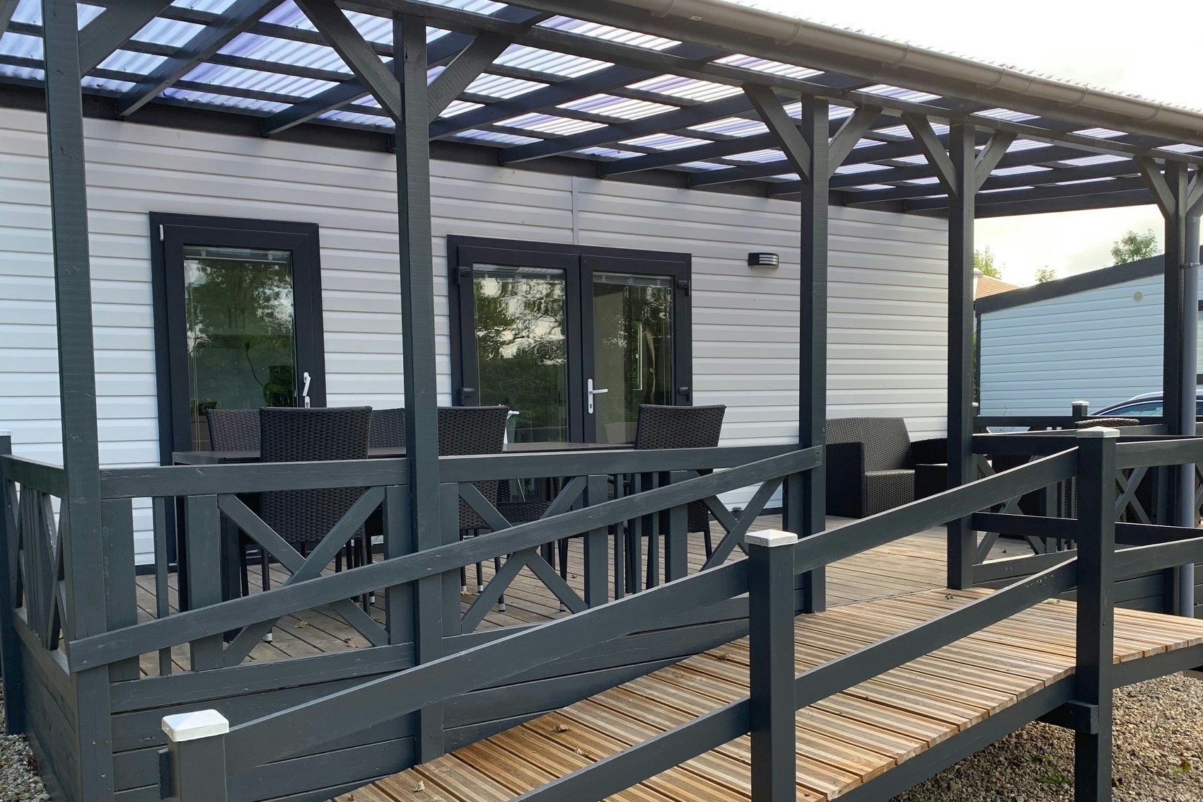Holzterrasse mit Geländer und überdachtem Bereich vor einem weißen Camping-Gebäude, mit Schotterboden und Sitzgelegenheiten.