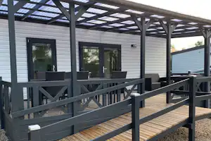 Holzterrasse mit Geländer und überdachtem Bereich vor einem weißen Camping-Gebäude, mit Schotterboden und Sitzgelegenheiten.