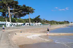 Wohnmobile am Strand hinter einer Natursteinmauer, Bäume und blauer Himmel, Menschen laufen am Ufer entlang.