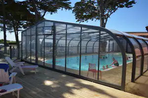 Überdachtes Schwimmbecken mit Glaswänden, Holzterrasse und Liegestühlen; im Wasser sind mehrere Personen zu sehen.