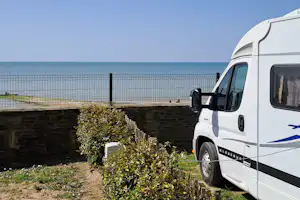 Weißes Wohnmobil am Campingplatz, Blick auf das Meer hinter einem Zaun und Hecke im Vordergrund.