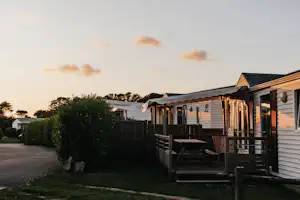 Campinghaus mit Terrasse und Markise, Holzgeländer und Fensterläden, im Hintergrund weitere Unterkünfte und Bäume.