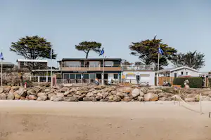 Campinggebäude mit Terrasse und Geländer, davor Bänke und Steine, im Hintergrund Bäume und blaue Fahnen.