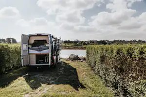 Offenes Wohnmobil mit Bett und Stauraum, Fahrräder am Heck, am Seeufer zwischen Büschen und Felsen.