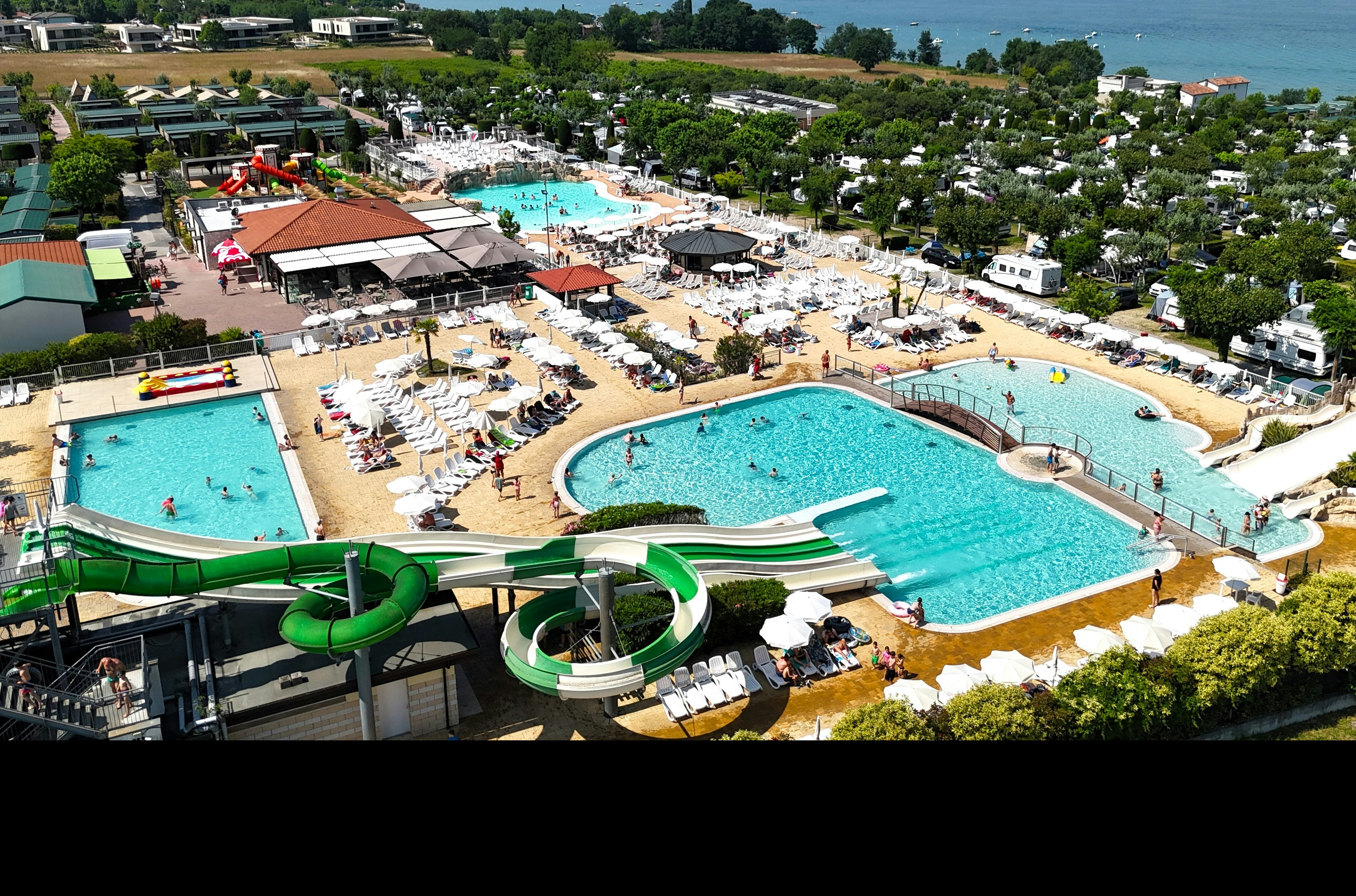 Luftaufnahme eines Campingplatzes mit mehreren Pools, Liegen und Sonnenschirmen sowie einem großen See im Hintergrund.