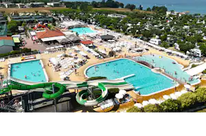 Luftaufnahme eines Campingplatzes mit mehreren Pools, Liegen und Sonnenschirmen sowie einem großen See im Hintergrund.